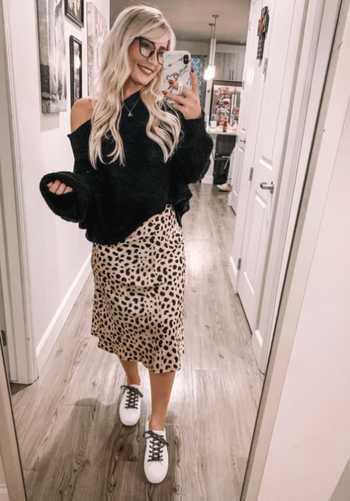 Reviewer in a black off-the-shoulder sweater paired with a leopard print skirt and sneakers, captured in a mirror selfie