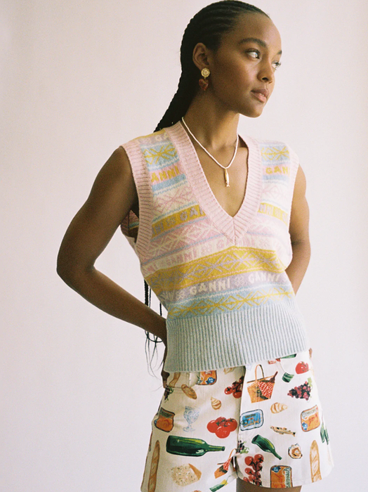The model is seen wearing a graphic V-neck sweater vest paired with patterned shorts.