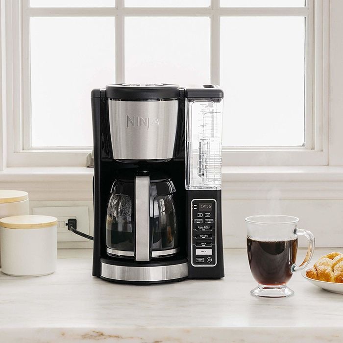 A lifestyle photo of the coffee maker sitting on a kitchen counter, next to a steaming cup of coffee.