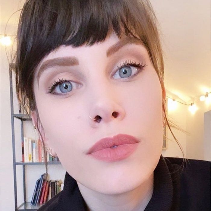 A reviewer wears a taupe-pink lipstick, demonstrating its soft, natural hue.