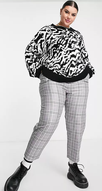 The model is sporting the same sweater paired with plaid trousers and sleek black boots.