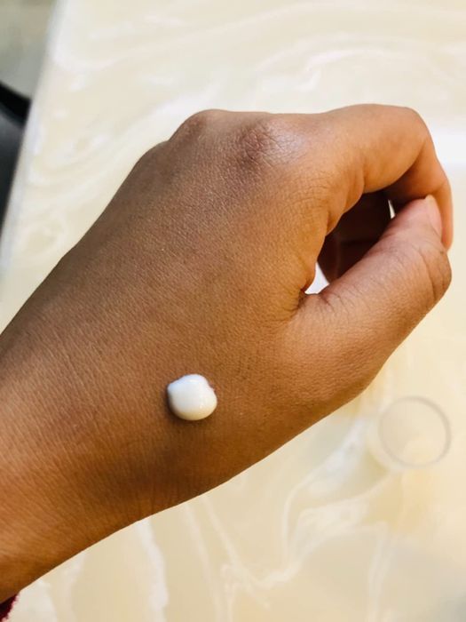 A reviewer with darker skin displays a white dot of sunscreen on their hand, emphasizing its blendable formula.