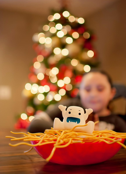 A child in the background with a Christmas tree, while the foreground features a bowl of noodles topped with a fun character-shaped topper.