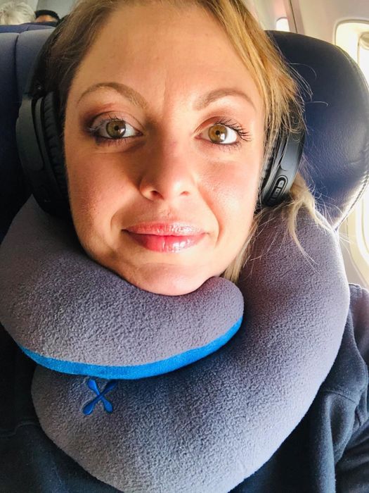 A reviewer wearing a gray wrap-around travel pillow while sitting comfortably in an airplane seat.