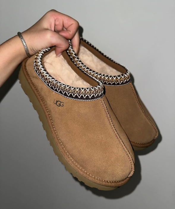 A reviewer showing off a pair of light brown slippers with a black and brown embroidered trim.