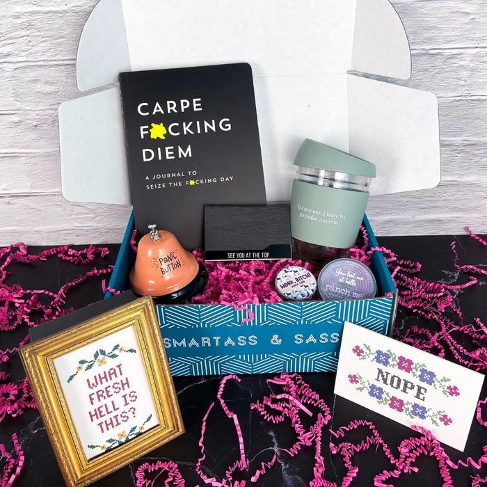 An open box filled with a framed cross-stitch, a reusable coffee cup, a bell, a book, and other fun items.