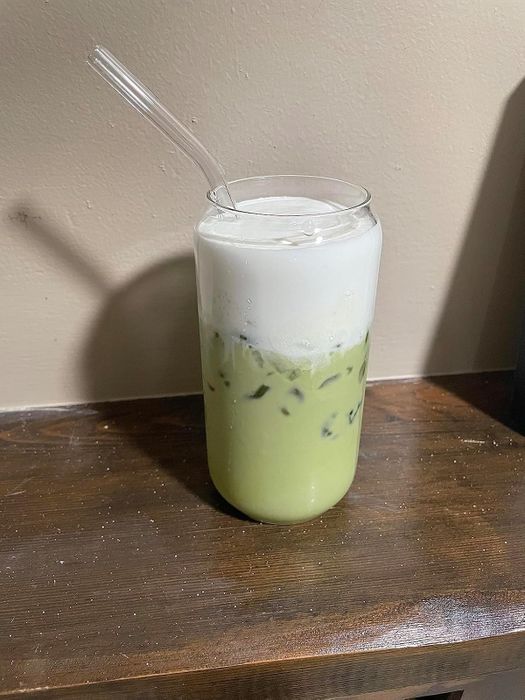 A glass of iced matcha latte topped with frothy milk foam, resting on a wooden surface