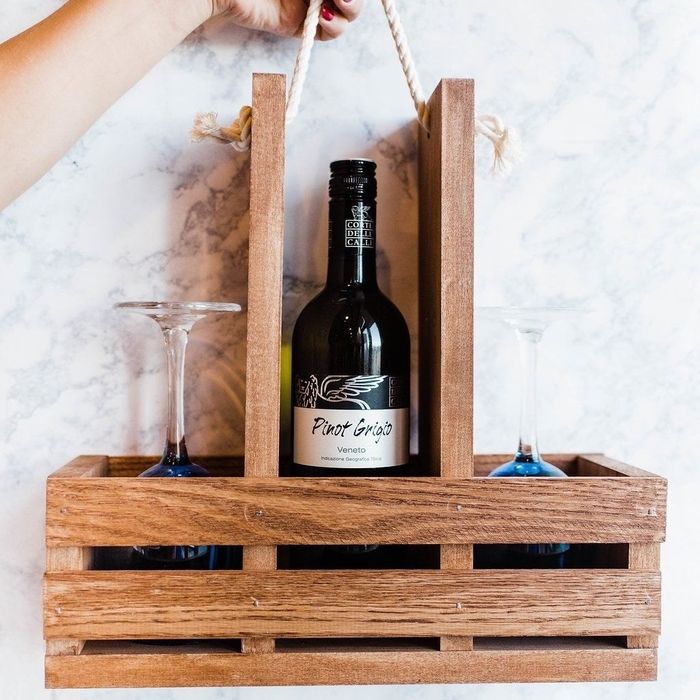 A hand holding a wooden crate with multiple compartments, featuring a top handle, filled with a bottle of wine and two glasses.