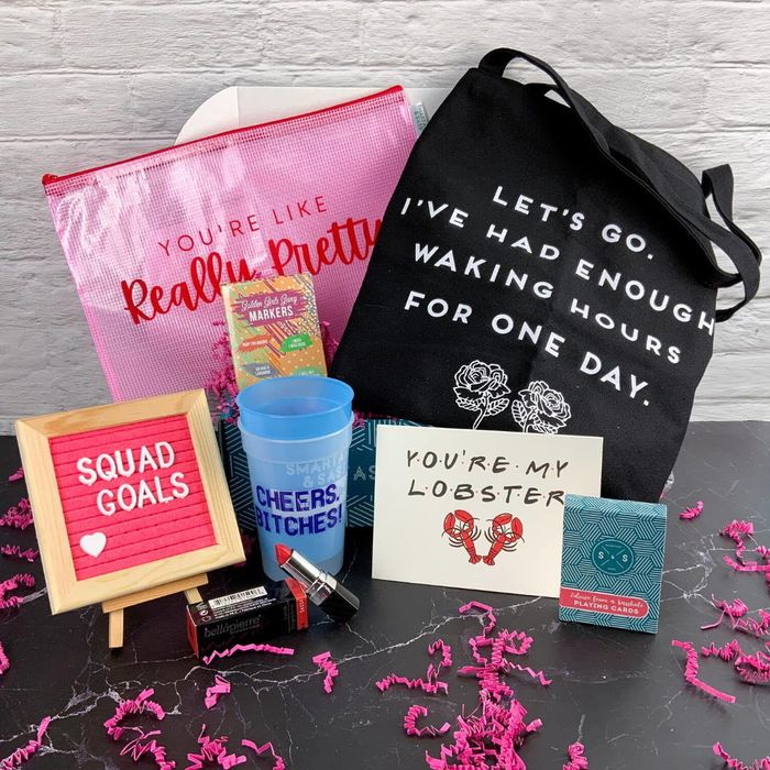 An open box containing a tote bag, card, cup, lipstick, message board, and additional small items.