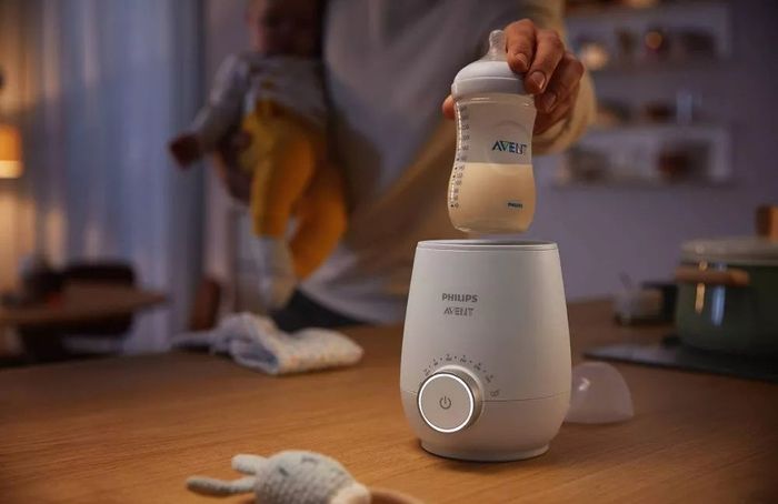 A hand places a baby bottle filled with milk into a white bottle warmer device.