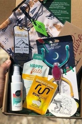 An open box containing a sticker, gum, a printed pouch, and an assortment of hiking and camping gear.