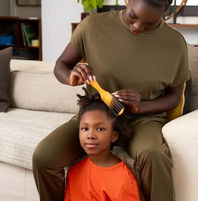 A woman gently combs a child's hair on a couch, both dressed casually, in a cozy home environment.