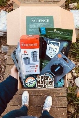 A top-down view of a hand presenting an open box filled with stainless steel cups, Adventure Blam, coffee, and various other outdoor and camping essentials.