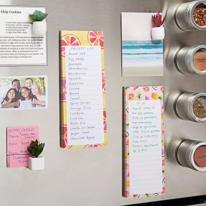 A magnetic fridge notepad with a grocery list, sticky notes with various lists, and a photo, all stuck to a refrigerator door.