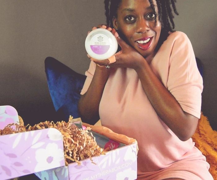 A reviewer sitting with an open subscription box on their lap, smiling, and holding a round tin of one of the products inside.