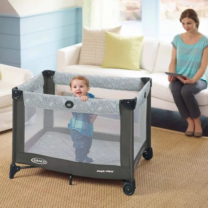 A toddler standing inside a gray and black pack and play, surrounded by mesh sides.