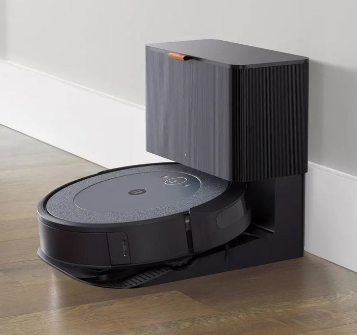 A robot vacuum cleaner is parked in its charging station, nestled against a wall on a hardwood floor.