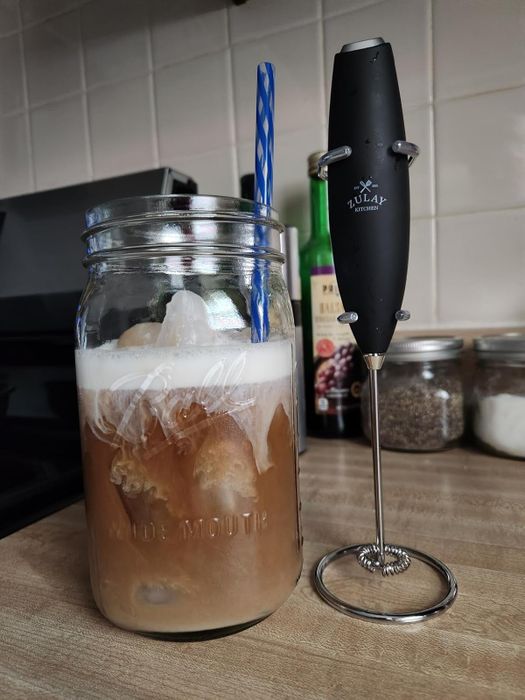 A mason jar filled with iced coffee and a blue straw, placed next to a black handheld milk frother on a kitchen counter