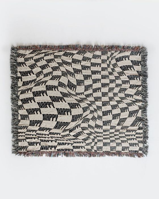 The throw blanket, in a soft cream shade, features black fringe trim along the edges with the word 'Nappy' repeated throughout the fabric.