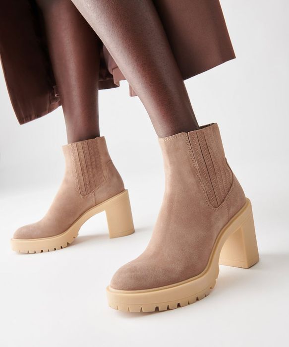 A person steps out in tan suede ankle boots with chunky heels and ribbed side panels, showing off a trendy and practical look.