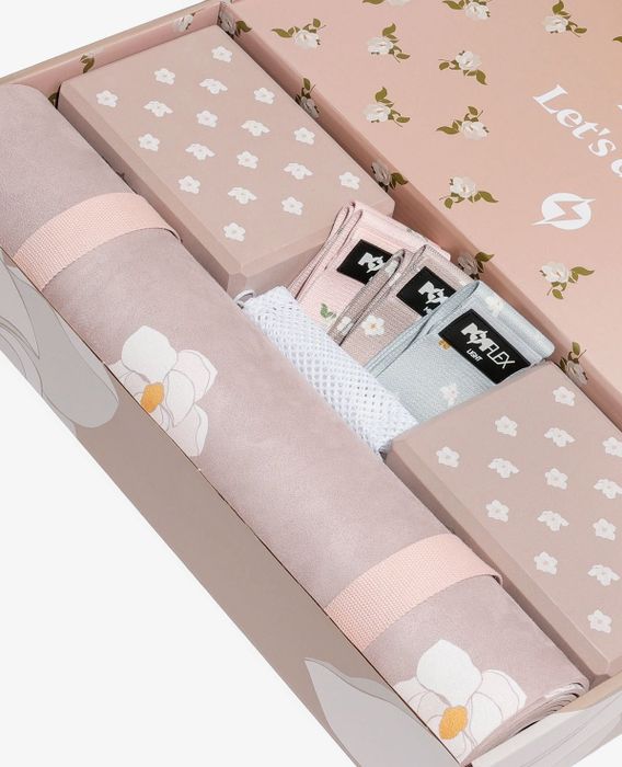 A soft pink box containing a yoga mat, two yoga blocks, three resistance bands, and a mesh bag, all beautifully adorned with magnolia flower designs.