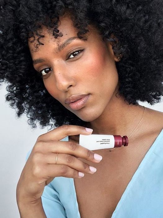The model is holding the small tube with a dark purple end, applying the product to their cheeks.