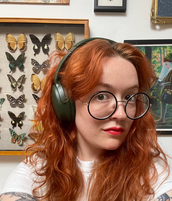 Me wearing large round glasses and headphones, standing in front of a wall decorated with framed butterfly displays and artwork.