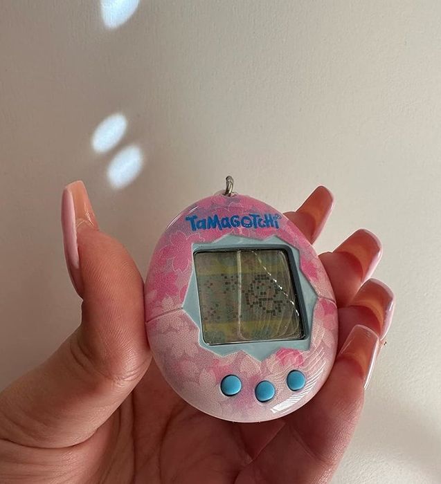 A hand holding a Tamagotchi, the iconic handheld digital pet, with a simple backdrop. The device features a small screen and buttons.