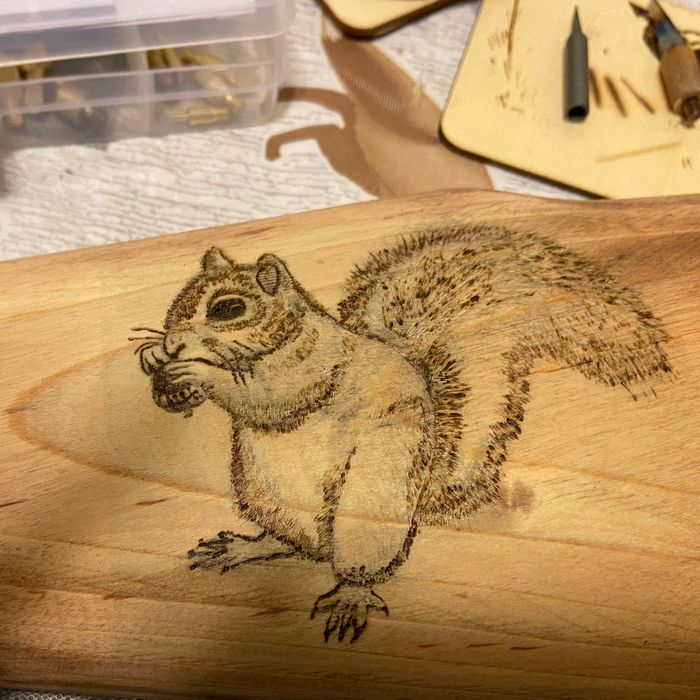 A wooden object featuring an intricate squirrel engraving, with tools and materials scattered in the background, hinting at a DIY or crafting project.