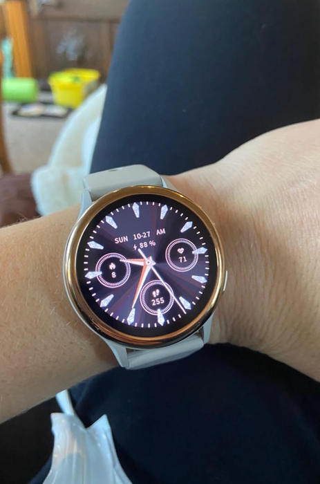 A person wearing a smartwatch, showing the time, date, battery level, and various fitness metrics. The watch has a light-colored band.