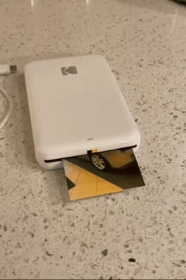 A compact Kodak photo printer producing a printout on a countertop.