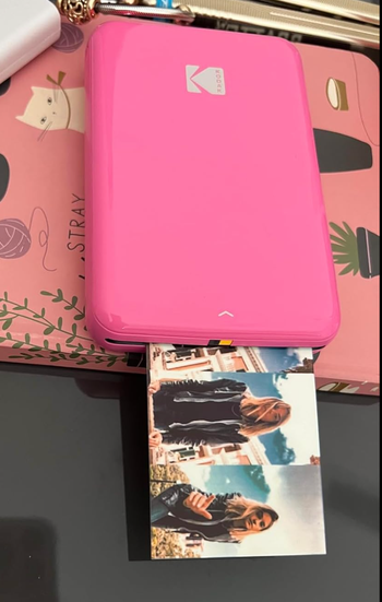 A portable printer producing a photo of two women outdoors, one holding a cup, with the printer placed on a pink book adorned with illustrations.