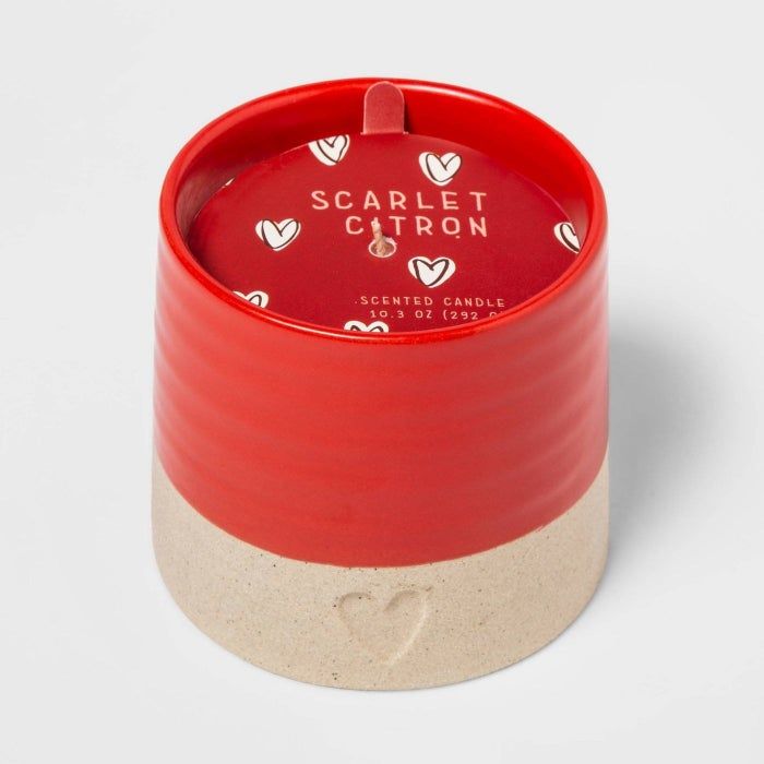 The candle features a circular ceramic holder with a red top and beige bottom, adorned with a small heart design on the base.