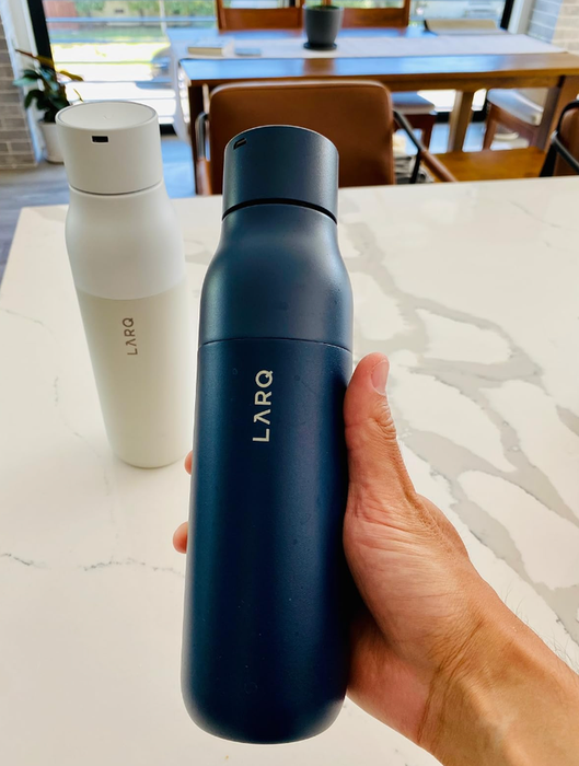 A hand holding a sleek dark LARQ water bottle on a marble table, with another bottle visible in the background.