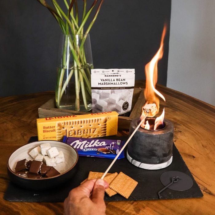 A s'mores kit complete with marshmallows, chocolate, and biscuits on a table; a marshmallow is roasting over a small flame.