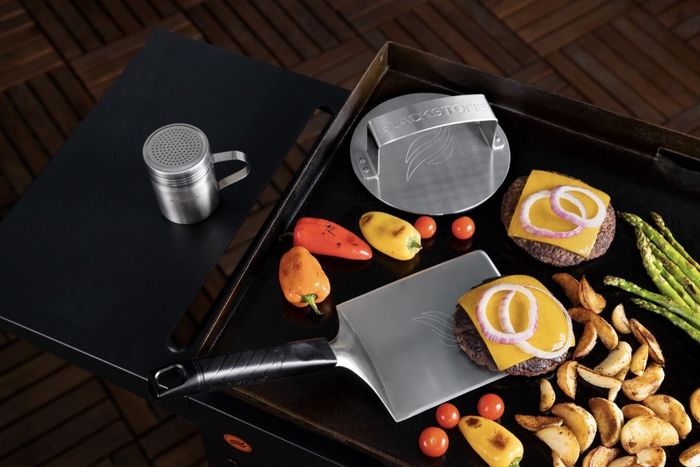 The three stainless steel tools are laid out on a grill beside burgers and a variety of vegetables.