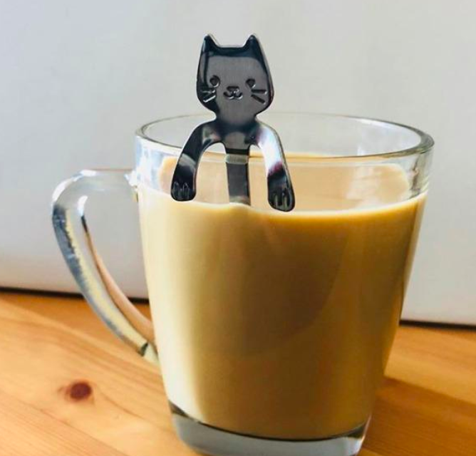 A cat-shaped spoon perched on the rim of a clear coffee cup.