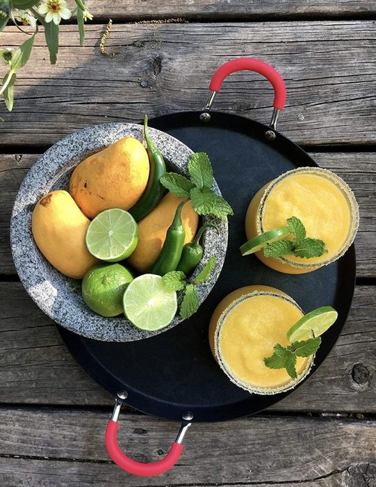 The mortar is crafted from grey and white granite, filled with limes, mangoes, and jalapeños, accompanied by two juices on the side.