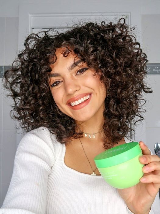 person with curly hair holding a green jar of hair mask