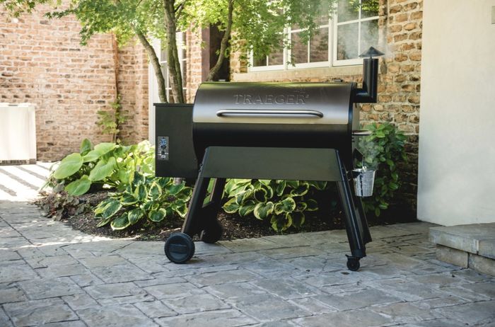 The taupe-colored Traeger oven, with its sleek black base and legs, is placed outdoors with smoke gently rising from the top.