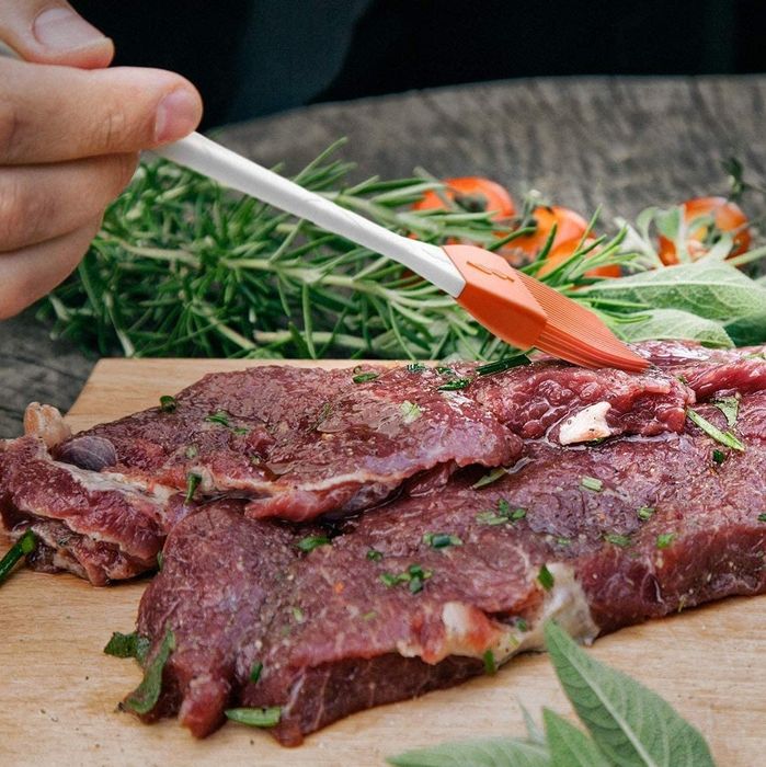 A red silicone basting brush applying a flavorful marinade to a piece of raw meat.
