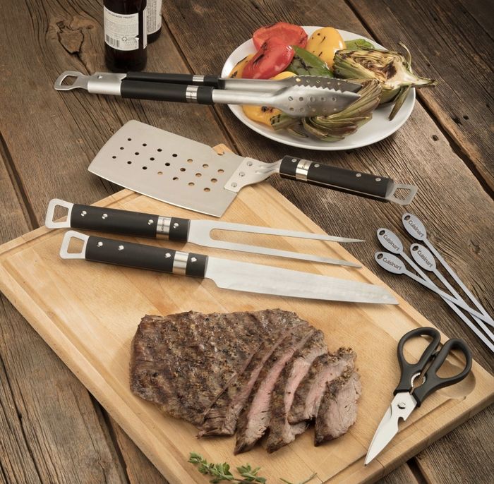 A bamboo cutting board with a slice of meat on top, surrounded by various metal and black grilling tools.