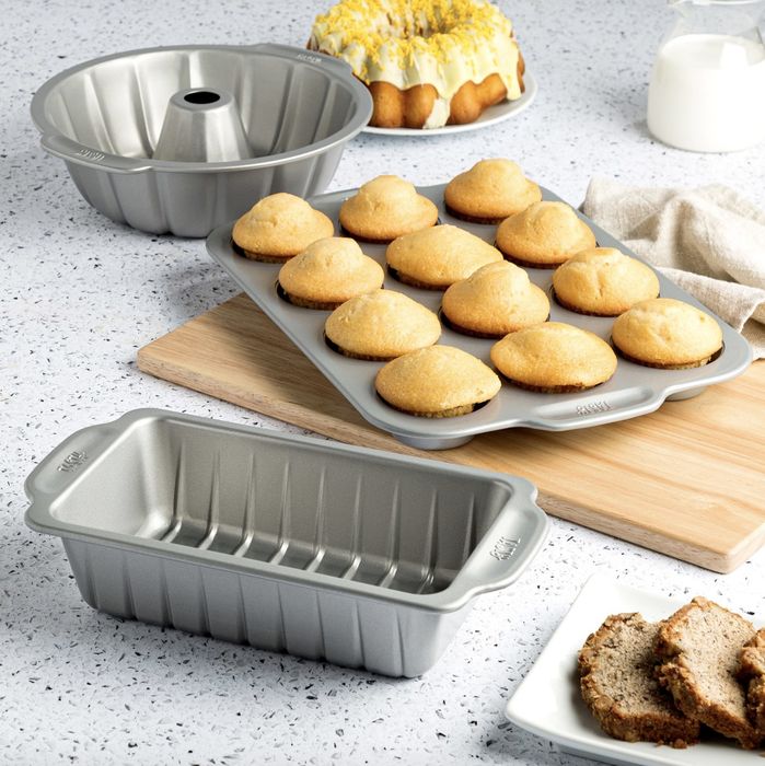 The set is made of stainless steel and is surrounded by an assortment of freshly baked muffins, a cake, milk, and bread.