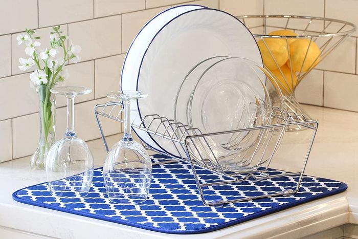 A dish drying rack loaded with dishes and topped with two wine glasses, all placed on a drying mat with a blue and white trellis design.