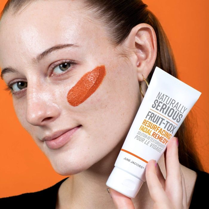 A person gently applies Naturally Serious Fruit-Tox Resurfacing Facial Remedy to their cheek, holding the tube close to their face.