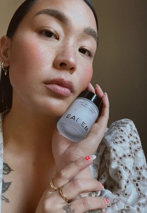 A person with a collarbone tattoo holds a jar of Eadem Cloud Cushion moisturizer near their face, dressed in a white lace outfit and adorned with rings and nail art.