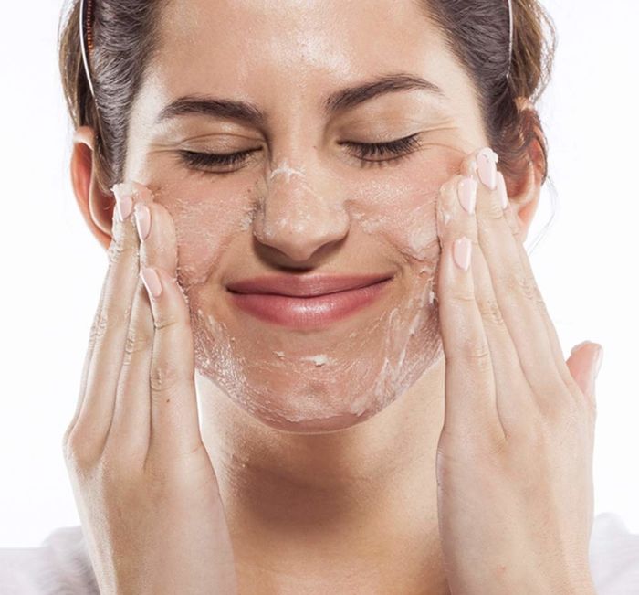 An individual is joyfully smiling with their eyes shut, gently applying a skincare product to their face using both hands.