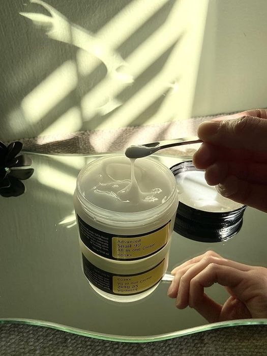 A hand is shown using a spatula to scoop out some 'Advanced Snail 92 All in One Cream' from a jar placed on a shiny surface.