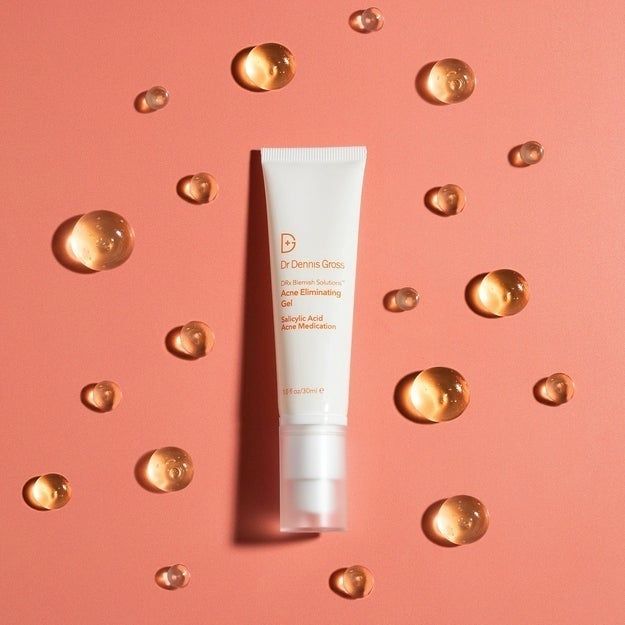A tube of Dr. Dennis Gross's Acne Eliminating Gel is displayed on a peach-colored background, with small droplets of the gel scattered around it.