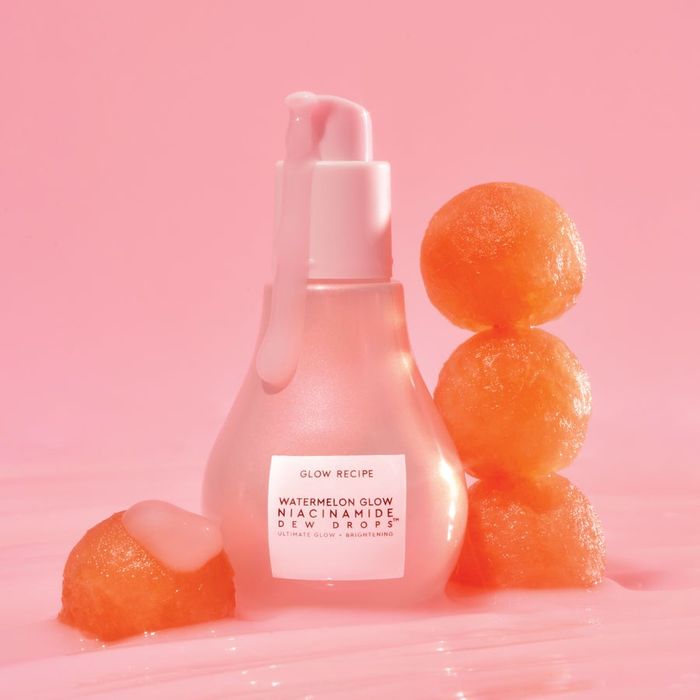Glow Recipe's Watermelon Glow Niacinamide Dew Drops bottle is showcased alongside watermelon slices on a soft pastel backdrop. The pump-top bottle displays a light pink serum inside.
